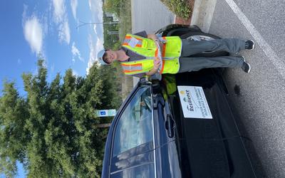 Jennifer Helgeson, holding a clipboard and wearing a high-vis vest stands next to a car. The car has a large sign on the side that reads “Center for Community Resilience Planning. Lumberton, NC Flood Study Team.” 