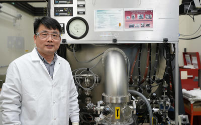 A researcher in a lab coat and safety glasses poses next to a large piece of lab equipment.