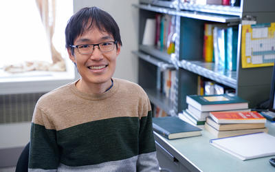 Leroy Jia poses smiling at his desk, with piles of books in the background. 