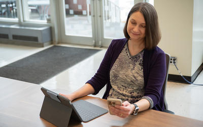 Julie Haney sits at a table in the NIST library with a laptop, looking at a smartphone in her hand. 