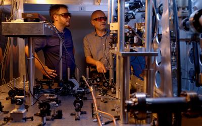 Two researchers in safety glasses stand behind a table holding various electronic and laser devices. A metal structure in the foreground is part of the atomic clock. 