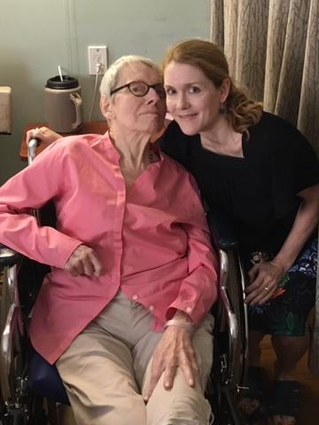 An elderly woman sits in a wheelchair next to a younger woman within a nursing home