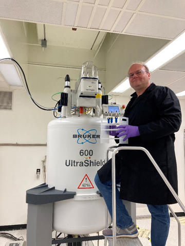 Aaron Urbas in a black gown and purple gloves on a stepstool in front of a large tank.
