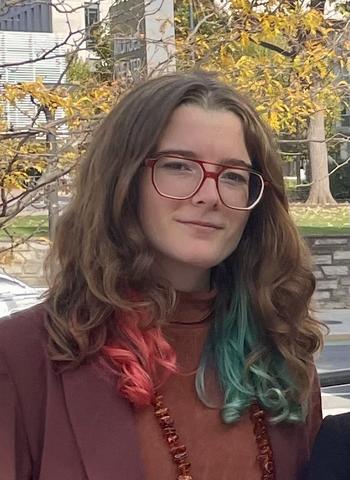 Headshot of Katarina Goodge. They have long brown hair, with some red on one side and green on another side. They are wearing glasses and a red top, standing outside, giving a soft smile for the photo.