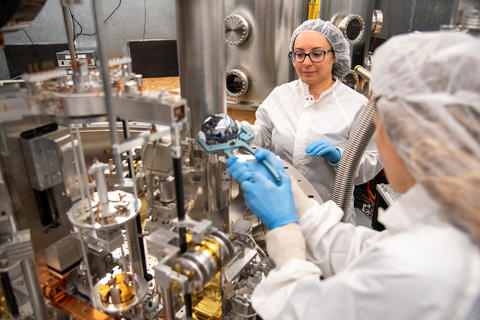 A researcher wearing a lab coat uses tongs to place a silvery sphere into a large scientific device as another researcher looks on. 