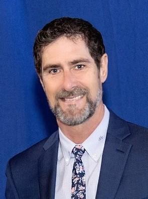 Headshot of a man with dark hair, beard and mustache wearing a suit and tie.
