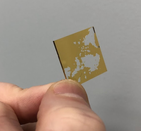 A tiny flat square with a chipped coating of gold is held between a finger and thumb. 