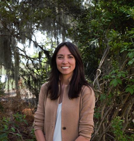 A woman with shoulder length black hair wearing a tan blazer and white shirt standing in front of a marshland.