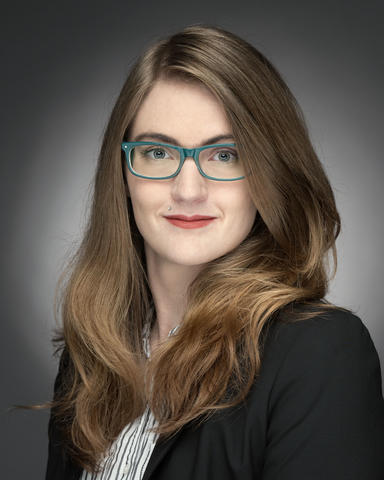 Indoor studio headshot of a woman with long, dark blonde hair in professional attire and glasses on a soft grey background.
