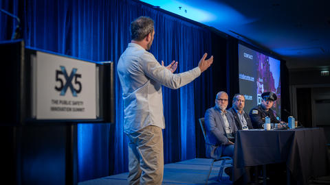 Presenter on stage in front of panel of judges at 5x5