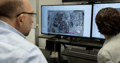 View from behind shows two seated researchers looking at a computer screen that shows an aerial view of a large outdoor site. 