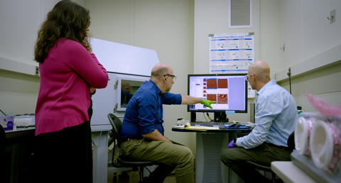 A man points to images on a computer screen in a lab as two other researchers look on, one seated and one standing. 