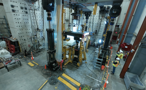 A high-ceilinged lab space is filled with large pieces of equipment, including a square structure in the center. A few people in hard hats stand at the edge of the room. 