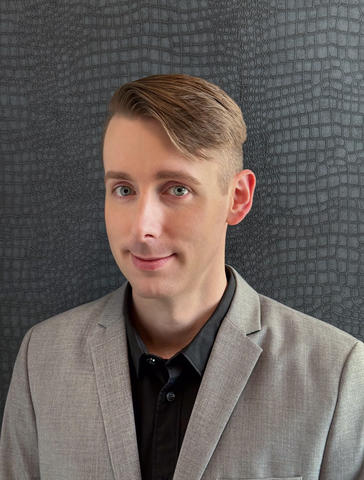 Headshot of Kyle Anderson, a person with short blonde comb over hair, wearing a black collared shirt and a gray suit jacket, smiling