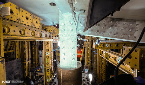 Heavy equipment and scaffolding surrounds a vertical concrete slab and attached horizontal beam, which is being cracked away.