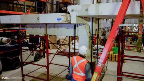 A person wearing construction safety gear looks up at a cracked concrete slab and beam arrangement held up by scaffolding in a large indoor space. 