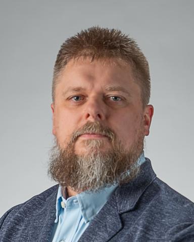 A professional portrait of a man with short, spiked brown hair and a full beard with hints of gray. He is wearing a light blue dress shirt under a textured dark blazer. He has a neutral expression and is facing the camera directly against a plain, light gray background.
