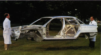 Two researchers in lab coats hold a sedan's aluminum frame off the ground in a grass field.