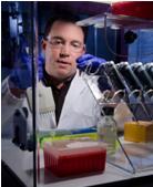 Kevin Kiesler wears a lab coat and goggles while working at a lab bench. 