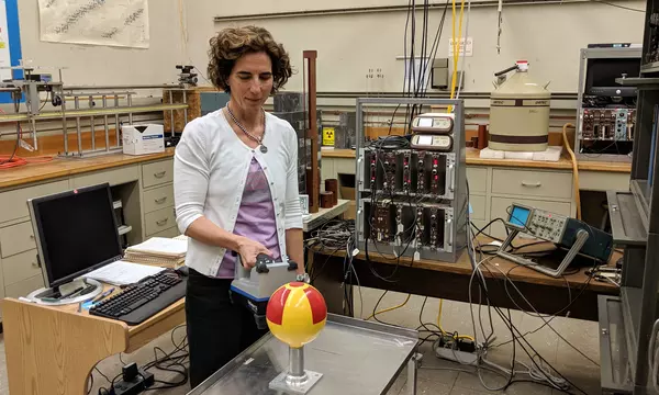 Woman standing in a lab holds a rectangular radiation detector near a metal globe that has been painted with a radiation symbol