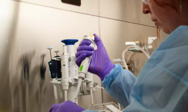 A woman in blue coveralls and purple gloves works at a lab table.