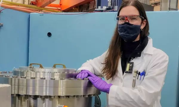 masked woman with glasses, purple gloves, and a labcoat stands next to a large aluminum sample chamber