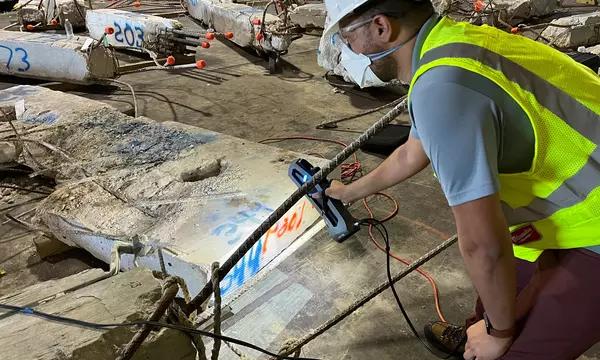 A researcher in safety gear leans over a large piece of concrete and rebar, pointing at it with a scanning device.