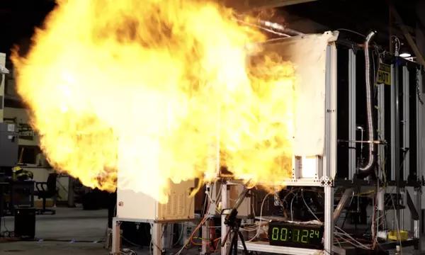 A fireball rushes out of the opening of a small metal compartment in a concrete room. 