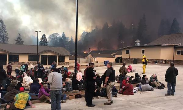 Groups of people sit or lie in a parking lot surrounded by low buildings as fires burn in the forest beyond.  