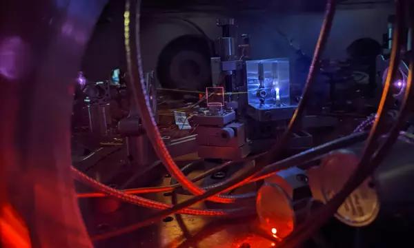 Table in darkened lab holds wiring and other laser electronics; a circular light source is situated above. 