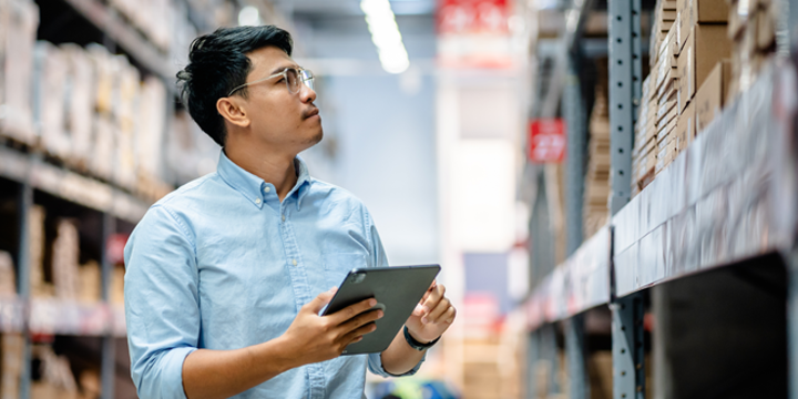 Man using a digital tablet to check the stock inventory on shelves in a large warehouse