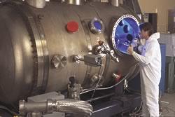 NIST physicist Charlie Tarrio stands in front of a machine that is long and silver like a sausage and has a blue light