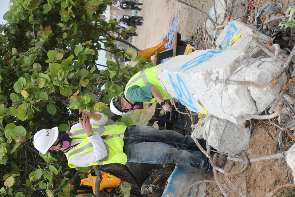 A man in a hard hat and other safety gear takes a picture of a broken concrete column with his phone.