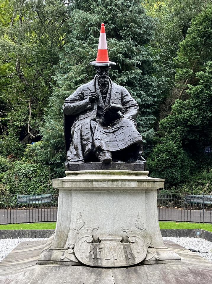 A statue of Lord Kelvin, seated with a notebook, stands in a leafy park. An orange traffic cone is placed on his head like a hat.  