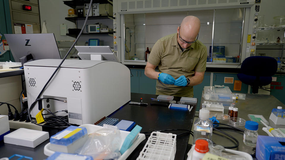 Thomas Forbes stands at a lab table near a white plastic 3D printer, looking down at materials he is preparing. 