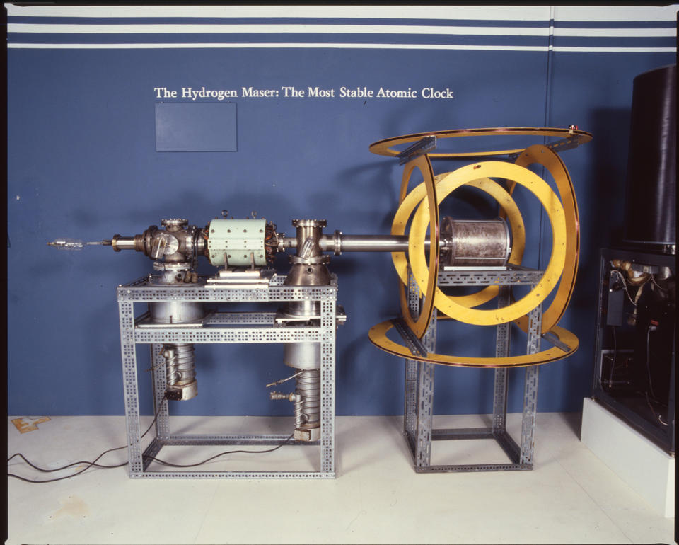 Museum display shows a cylindrical device lying horizontally on two mounts. Text on wall reads: The Hydrogen Maser: The Most Stable Atomic Clock. 