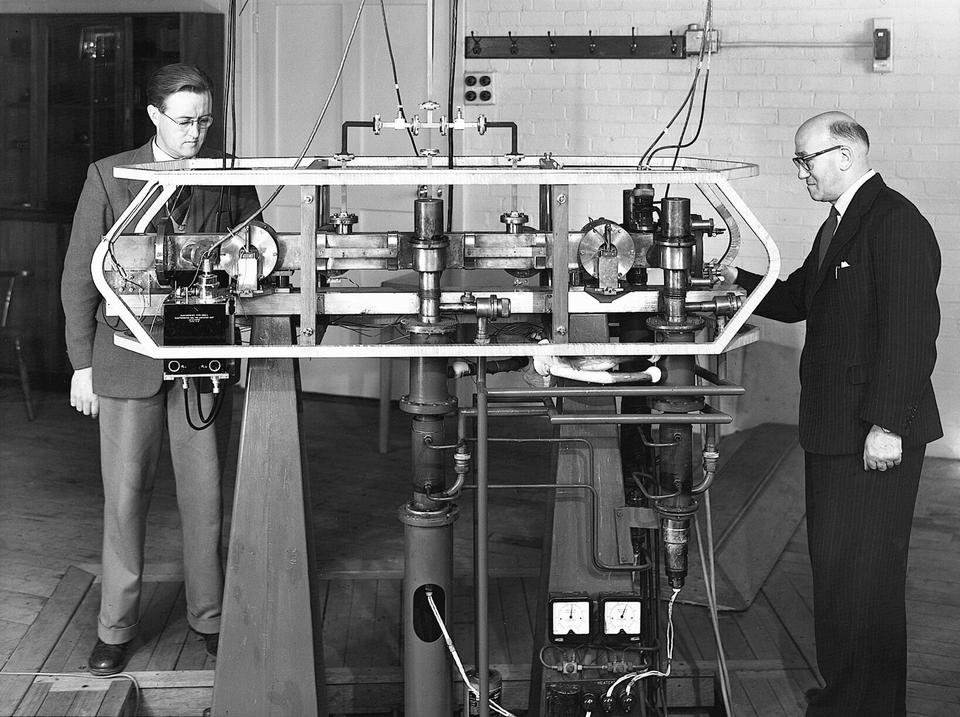 Historical photo shows two men standing at either end of a large device with a horizontal rectangle shape. 