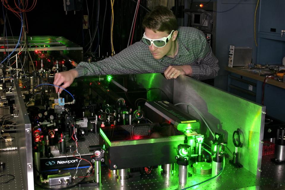 A researcher wearing safety glasses leans over a table crowded with laser devices in a darkened lab. 