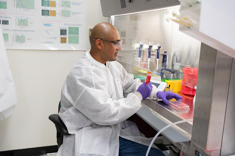 A researcher wearing safety gear sits at a lab table working with a tray of samples and a large vial holding red liquid.