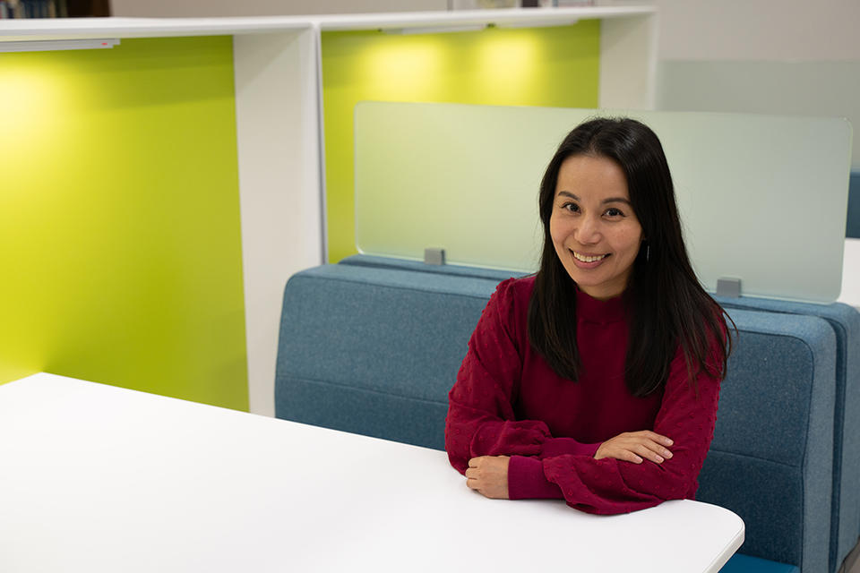 Mei Lee Ngan poses smiling and sitting at a white table with a bright green wall behind her. 