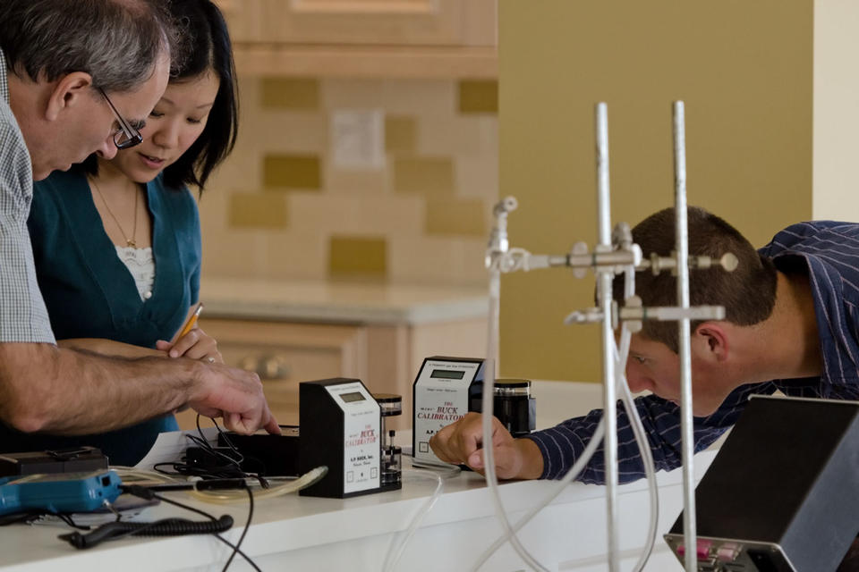 Three researchers stand and look down to examine various small electronics and wiring on a kitchen counter.