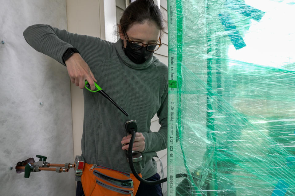 A researcher standing outside a house holds a lighter to the end of a hose-like device leading into an area closed off by plastic wrap. 