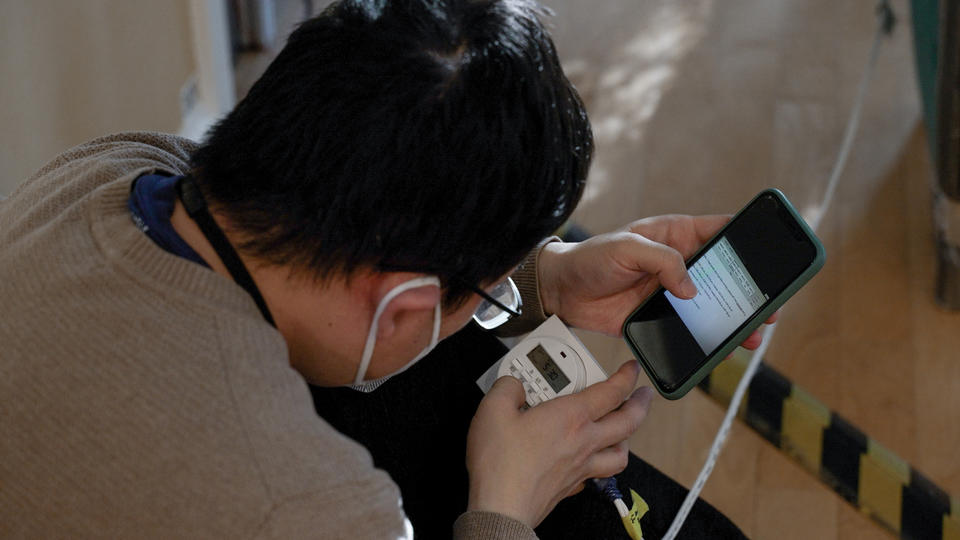 A researcher bends over a digital readout on a piece of equipment, comparing it to information on his phone. 