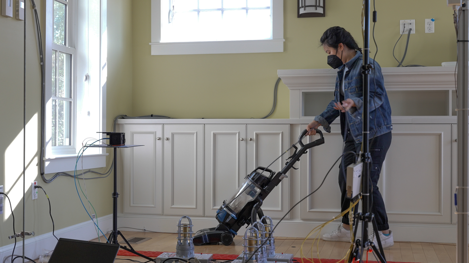 A researcher runs a vacuum cleaner over the wood floor in a living room that's empty except for scientific equipment and wiring. 