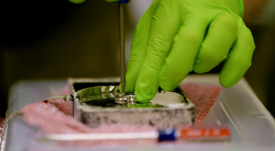 Close-up shows a hand in a green glove holding the center of a metal disk while using a pointed tool to remove a part. 