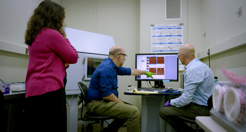 A man points to images on a computer screen in a lab as two other researchers look on, one seated and one standing. 