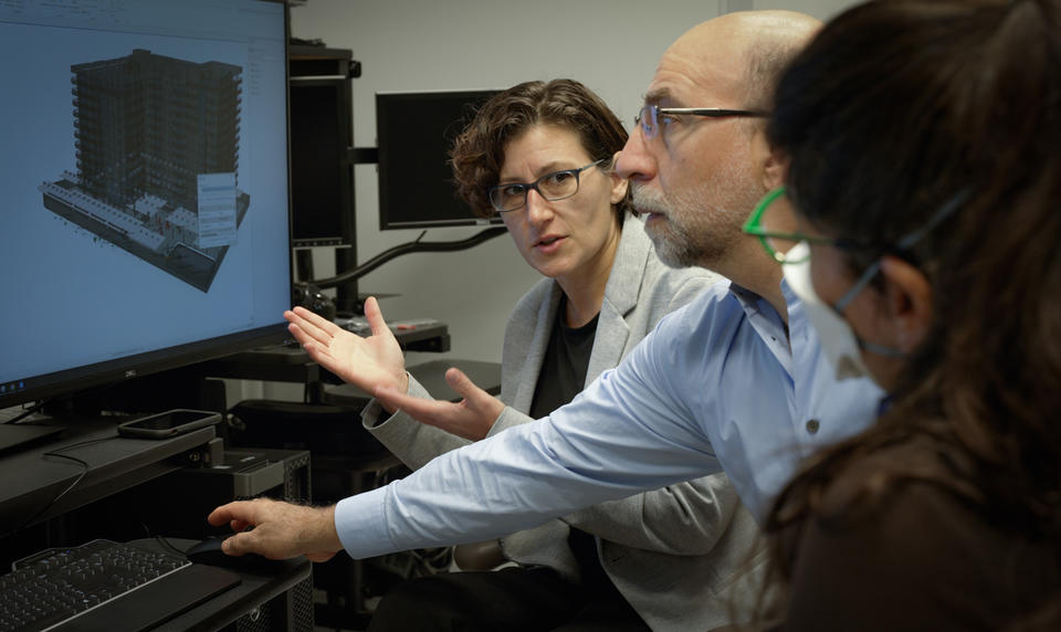 A woman gestures to a building model on a computer screen as she speaks to the two other researchers sitting near her. 