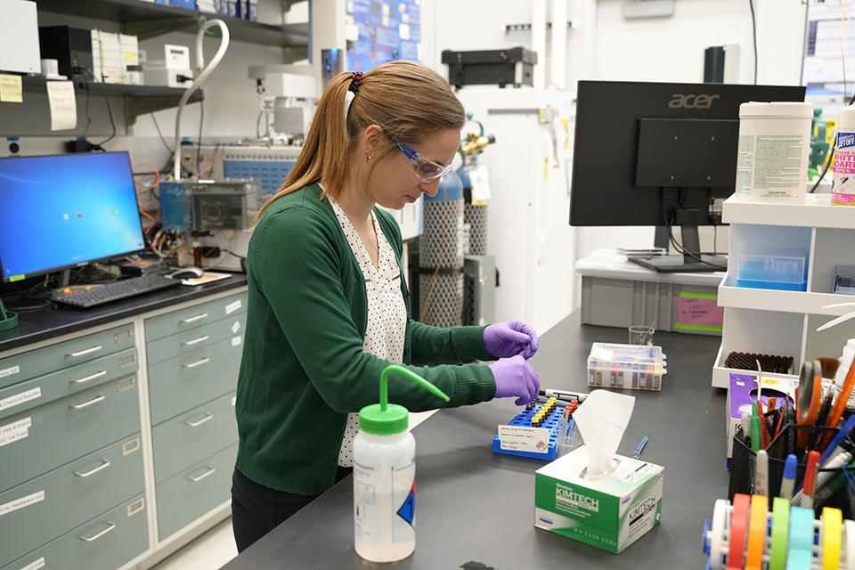 Briana Capistran wears safety glasses and purple gloves as she prepares small vials at a lab table. 