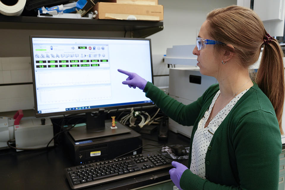 Briana Capistran wears safety glasses and purple gloves as she points to a computer screen showing information from lab analyses. 