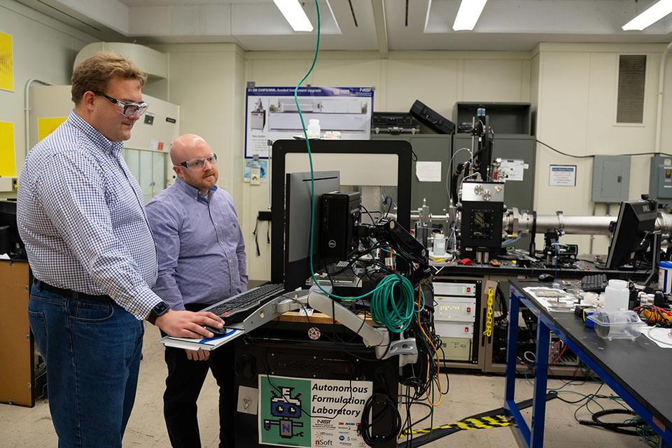 Two researchers in safety glasses stand looking at a computer screen in a lab filled with equipment.
